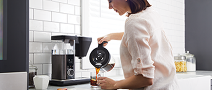 A woman pours herself a cup of coffee in front of a black coffee maker on a counter.