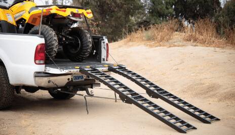 A pair of black foldable loading ramps positioned against the open tailgate of a white pickup truck. A yellow all-terrain vehicle is in the truck’s bed.