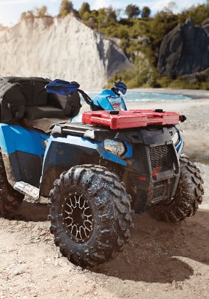 A blue-and-red ATV stands on the beach near a lake in a mountain setting.