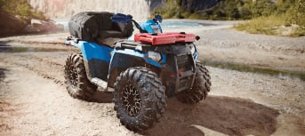 A blue-and-red ATV stands on the beach near a lake in a mountain setting.