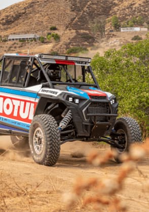 A blue-and-red UTV drives along a dirt path in a mountain setting.