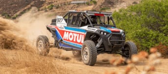 A blue-and-red UTV drives along a dirt path in a mountain setting.