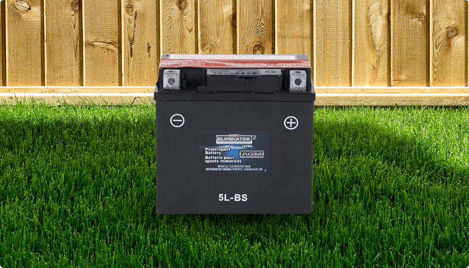 A black MOTOMASTER ELIMINATOR AGM Factory-Activated Powersports Battery rests on the grass near a wooden fence. 