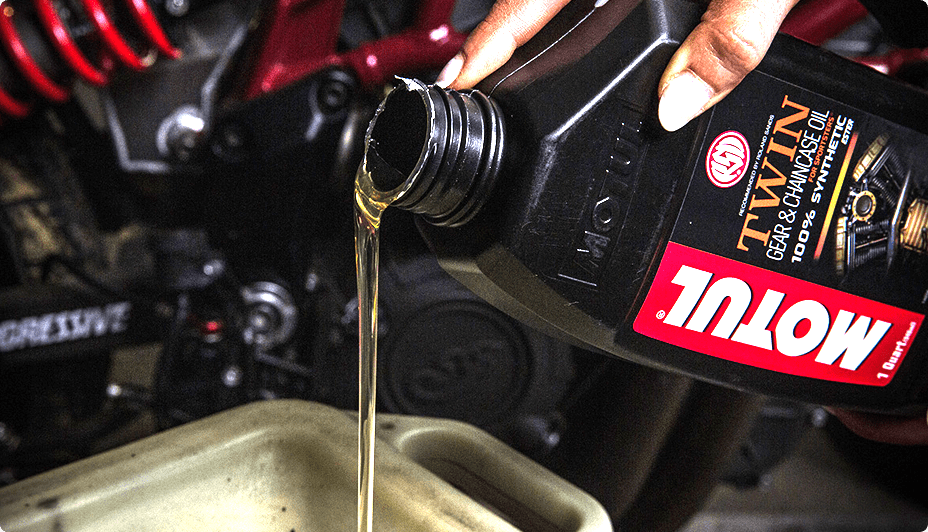 A hand pours a black bottle of Motul Twin 20W50 Synthetic Motorcycle Motor Oil into an oil pan. 