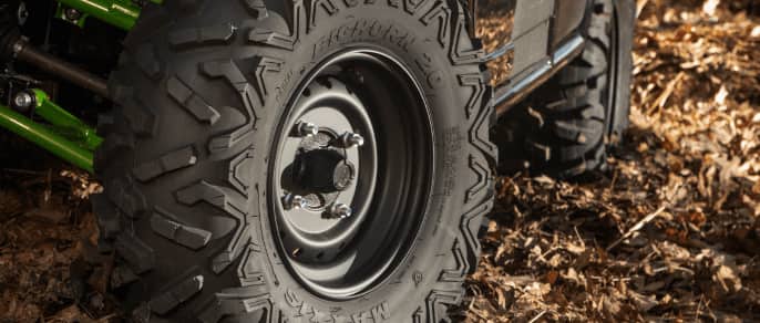 A close-up of an Maxxis ATV tire resting on ground covered in dry leaves.