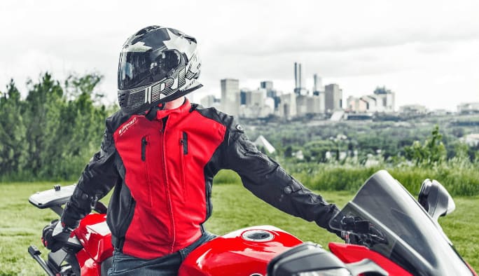 A motorcyclist in black-and-red motorcycle jacket and black-and-grey helmet with a tinted visor sits on a red motorcycle. A cityscape is visible in the distance.