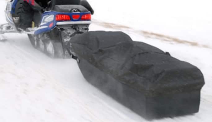 A blue snowmobile tows a black snowmobile toboggan along a snowy trail. A black cover is lashed across the toboggan.