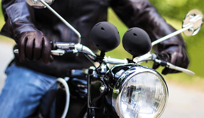 Deux haut-parleurs noirs montés sur le guidon d’une motocyclette. La motocyclette est pilotée par un conducteur portant des gants en cuir bruns et un manteau en cuir brun.