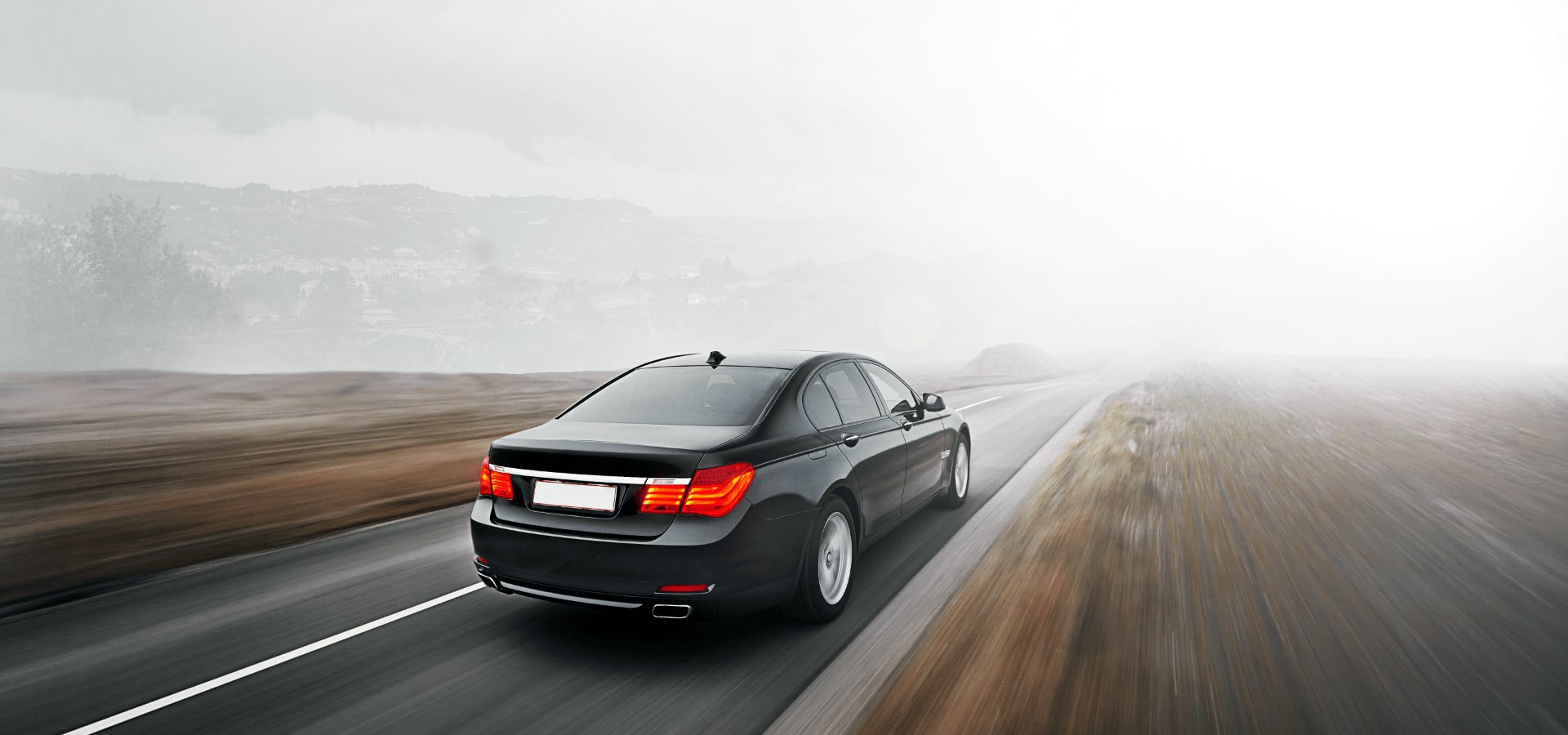 A black four-door sedan driving on a two-lane highway on a foggy day with mountains in the distance.