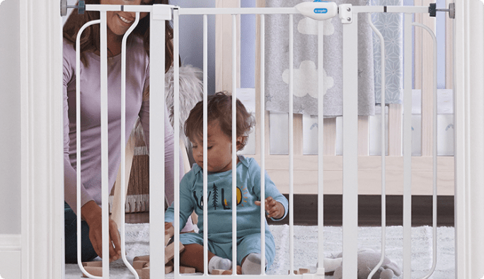 An adult plays with a toddler behind a white baby gate.