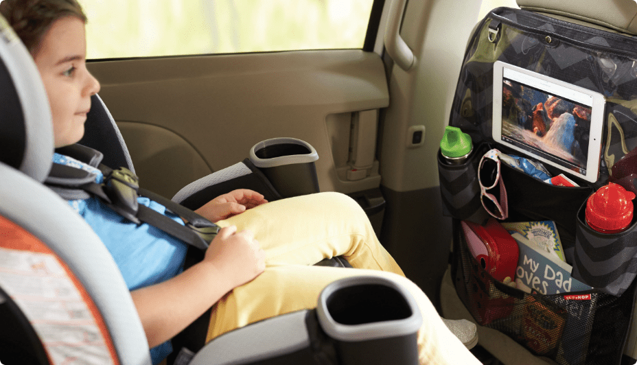 A child in a car seat looks at the screen of a tablet secured in a Skip Hop Style Driven Backseat Organizer attached to the back of a car’s driver’s seat.