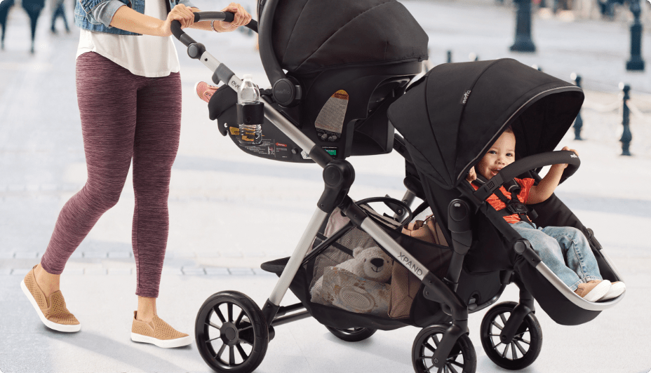 A toddler peeks out from under the canopy at the front of an Evenflo Pivot Xpand Modular Single to Double Travel System stroller pushed by an adult. Travel Systems