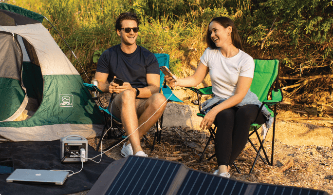 Two people sit on camping chairs in a sunny outdoor setting with their digital devices hooked up to a solar-powered battery.