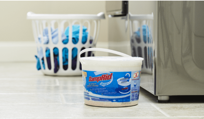 A 4-pound bucket of DampRid Fragrance-Free Hi-Capacity Moisture Absorber rests next to a washing machine and a laundry basket.