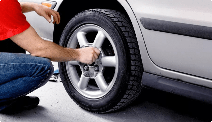 A crouching person loosens the lug nuts of a car’s tire by hand. 