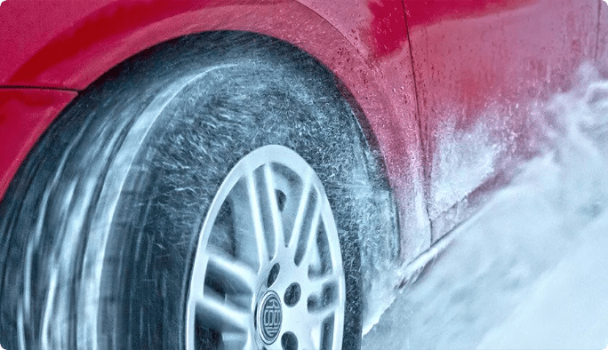 A close-up of the front tire of a red car driving through snow. 