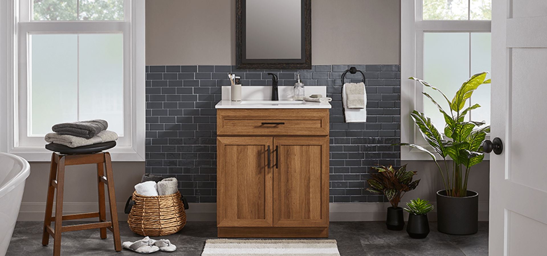 Bathroom with natural wood-finish vanity, dark wood stool, canvas cape basket, mirror with black frame.