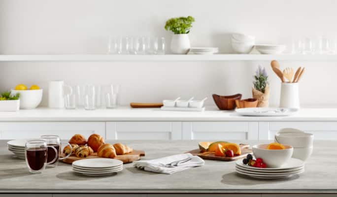 Étagères blanches dans la cuisine avec de la vaisselle, de la verrerie et des aliments pour le petit-déjeuner sur le plateau de la table.