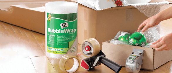 Person packing away Christmas decorations in a cardboard box with bubble wrap. Also pictured, a roll of packing tape, tape gun, roll of bubble wrap, and packing tape with dispenser.