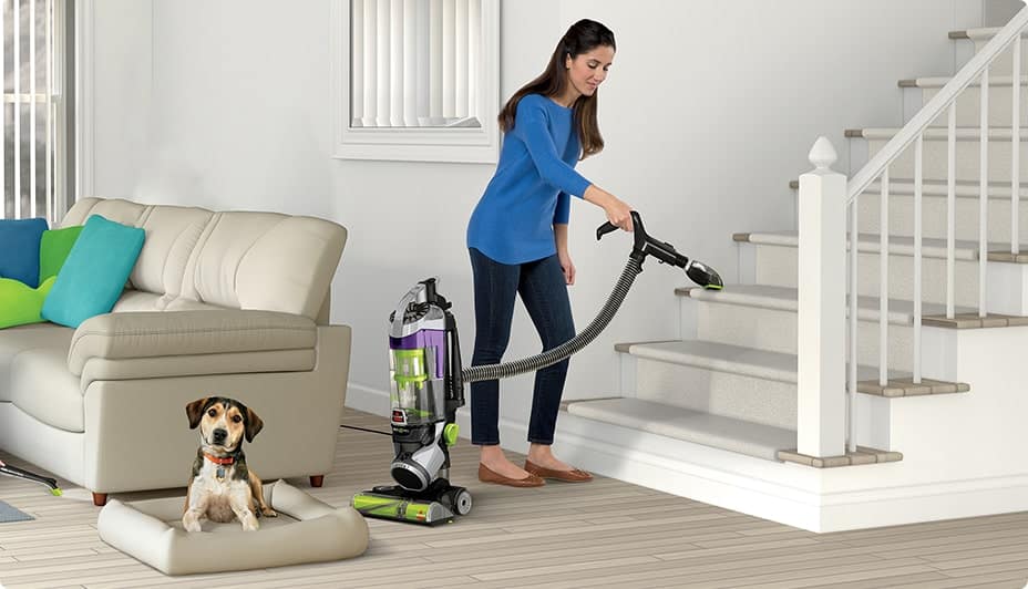 A woman vacuums the stairs with an upright vacuum and a dog sitting on a dog bed on a hardwood floor.