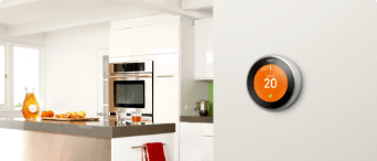 Smart thermostat device mounted to a wall. Around the corner is a modern-looking kitchen with various items on the counter.