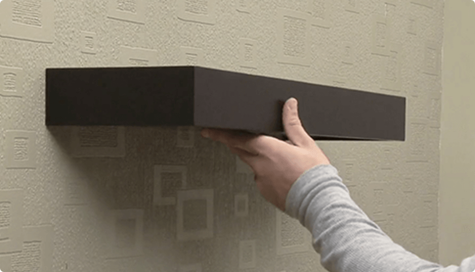 A hand holds a brown floating shelf against a wall.