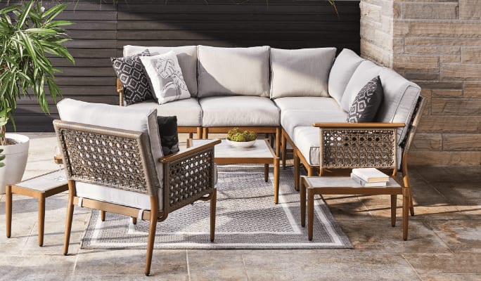 A wicker loveseat sectional and chair set with cream-coloured cushions in a backyard.