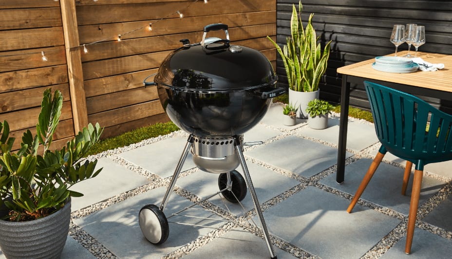 A black 22-inch Weber Master-Touch Portable Charcoal Grill set against horizontal-slat fencing in a backyard. 