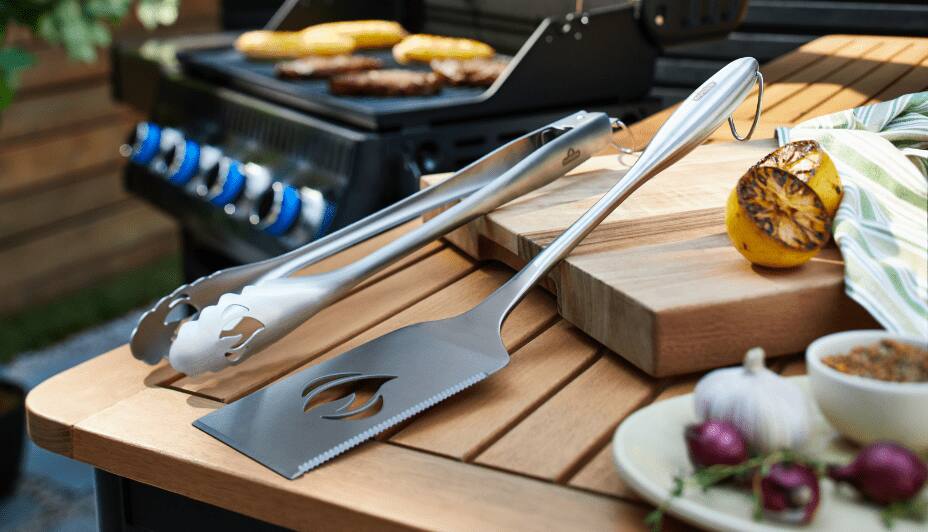 A stainless-steel spatula and set of tongs resting on a picnic table in a backyard.