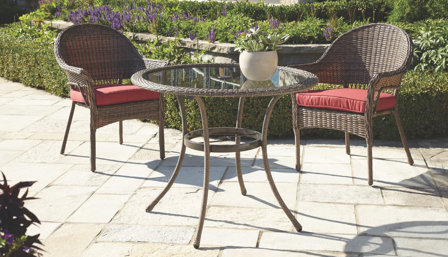 A round wicker outdoor table with a glass pane surface and two patio armchairs surrounding it.