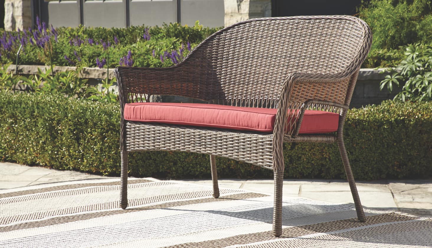 A brown wicker patio loveseat with a large red cushion sitting in sunlight. 