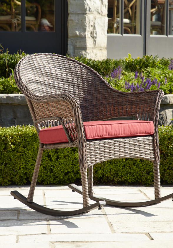 A brown wicker patio rocking chair with a red cushion placed on it sitting in sunlight.