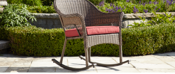  Un fauteuil de jardin berçant en osier brun avec un coussin rouge présenté dans un décor ensoleillé.