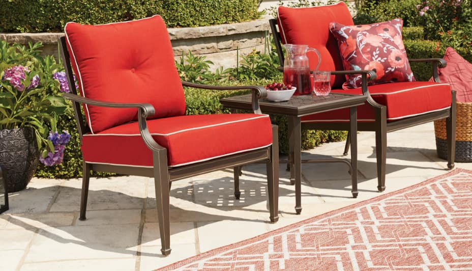 Two simple, elegant outdoor arm chairs with red cushions on each of their backs and seats, with a small metal table placed between them. 