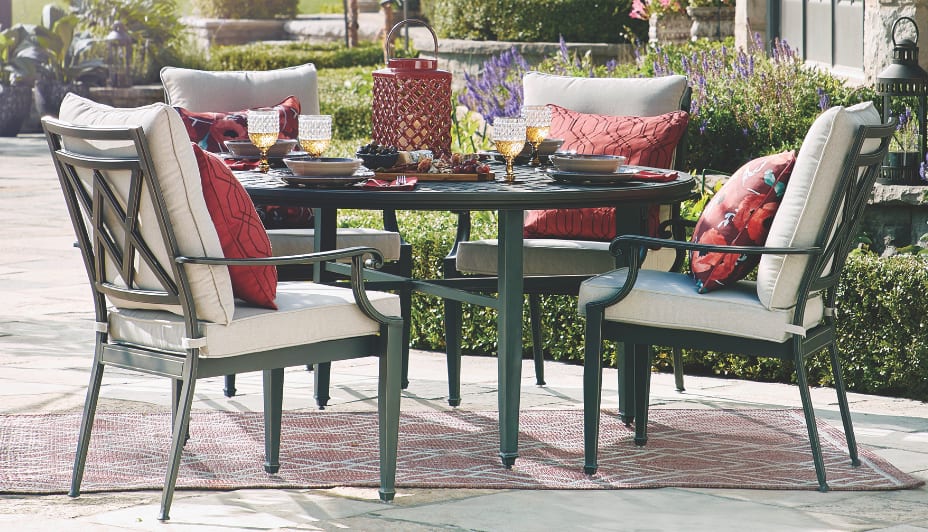  A large, circular outdoor dining table surrounded by four cushioned chairs in a backyard.