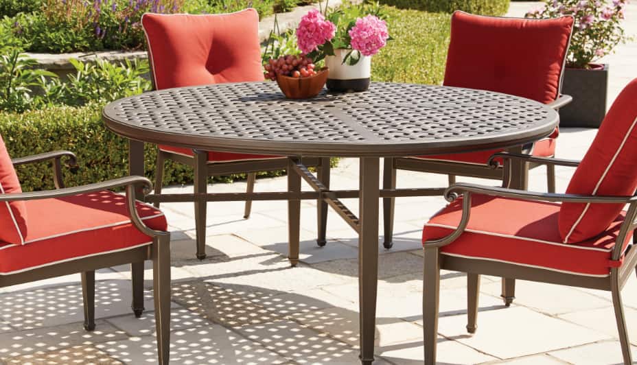 A large, circular, metal outdoor dining table surrounded by four red-cushioned chairs in a backyard.