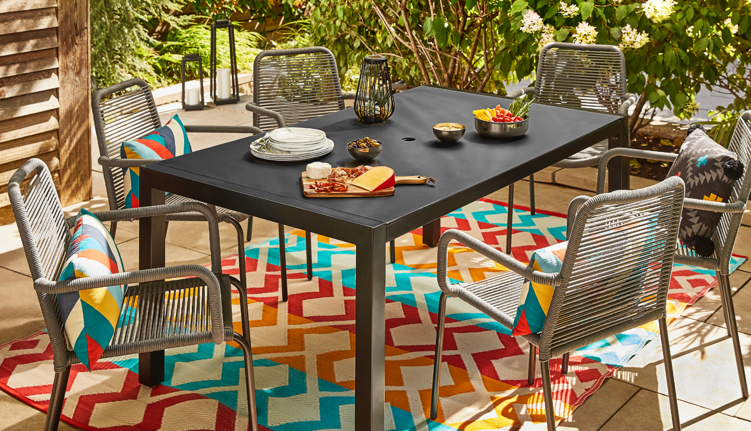 A black outdoor dining table with various foods and platters placed on top, with five grey wicker chairs surrounding it with some vibrant pillows on three of the them.