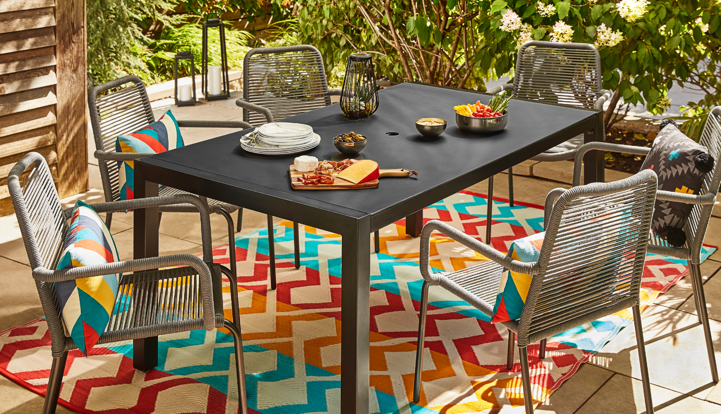 Une table à manger extérieure noire avec plusieurs plats et aliments sur le dessus et cinq chaises grises en osier avec des coussins vifs placés sur trois des chaises.