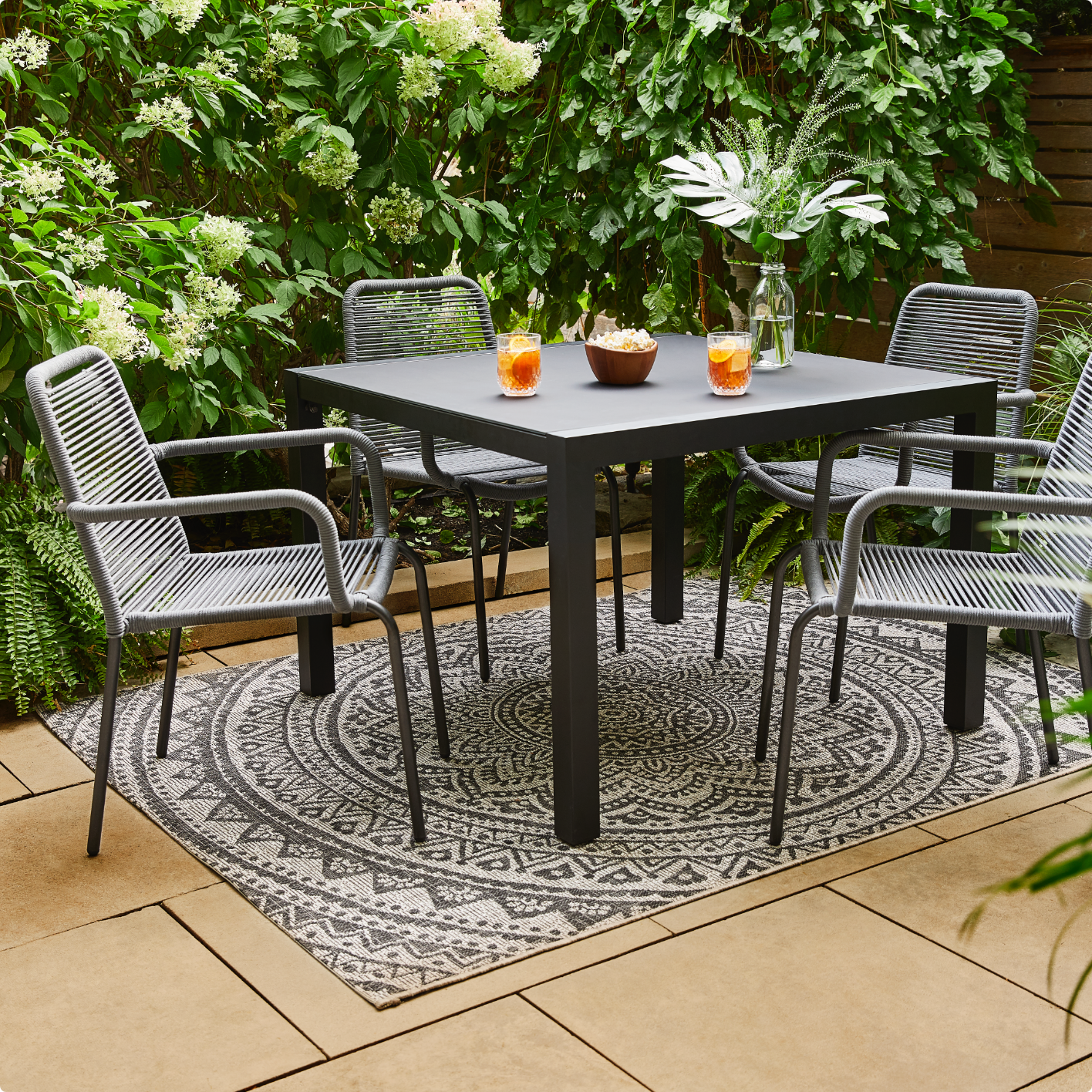 A smaller black outdoor dining table with some drinks on top, four grey wicker chairs surrounding it amidst a pleasant, secluded outdoor space. 