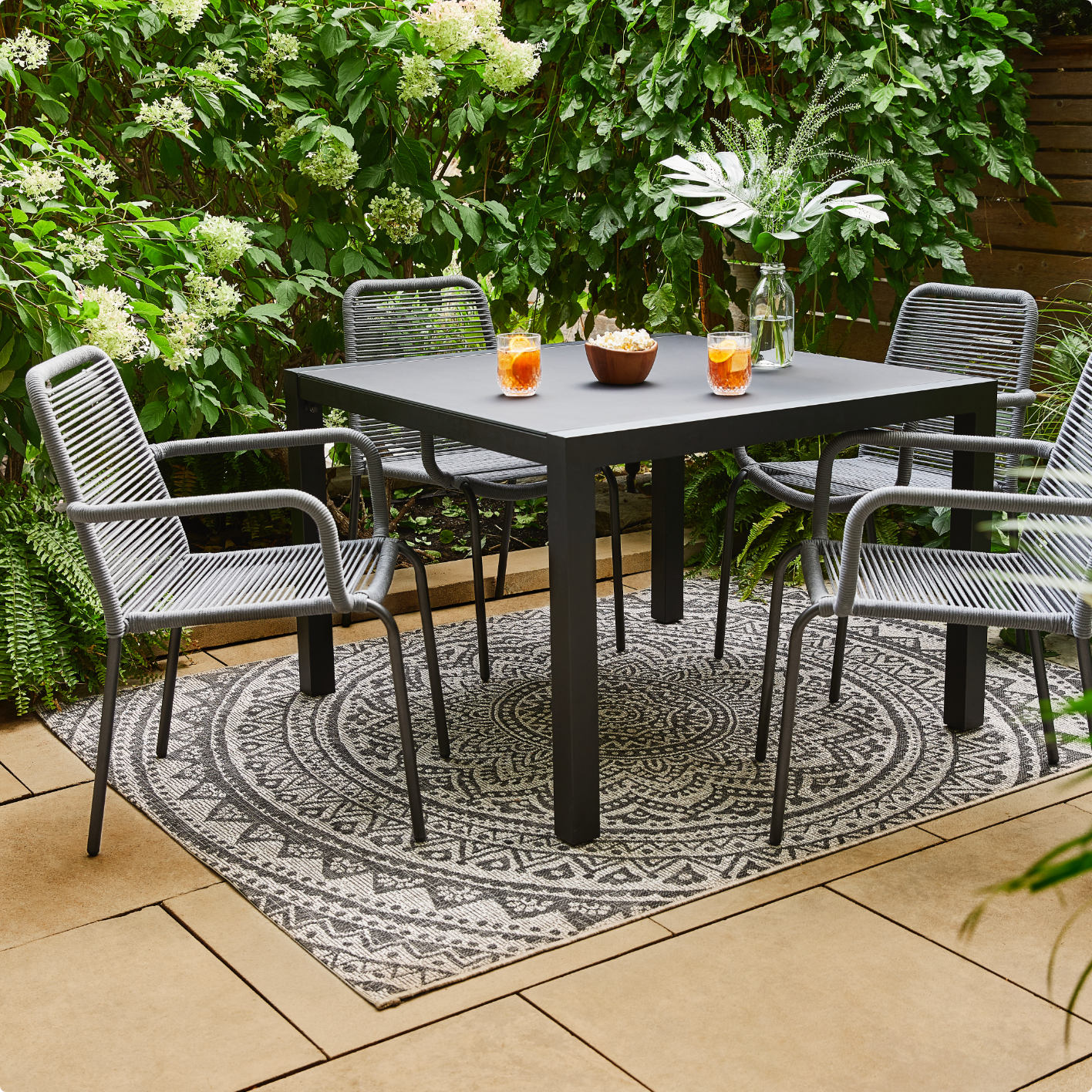 Une petite table à manger extérieure noire avec des boissons placées sur le dessus, quatre chaises grises en osier dans un espace extérieur. 