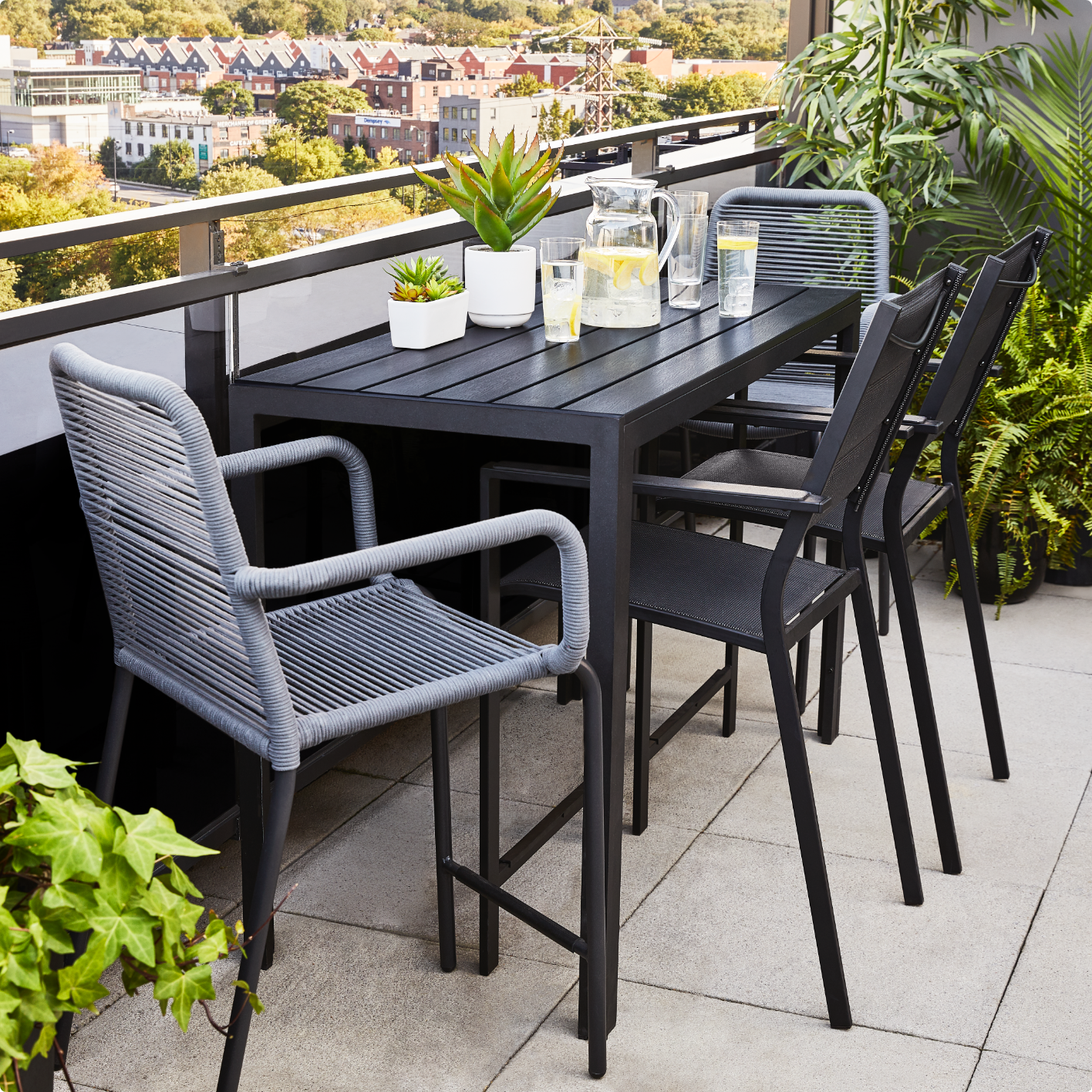 Une petite table à manger extérieure noire sur un balcon, quatre chaises grises en osier et des verres d’eau au citron placés sur la table. 