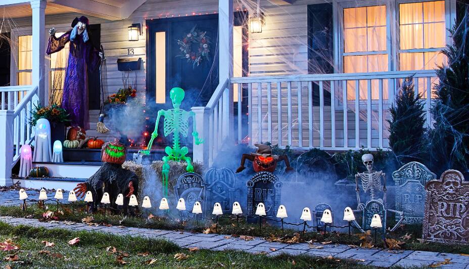 A standing witch and lit-up ghost figures, a skeleton, tombstones and ghost stake lights decorating the outside of a home for Halloween 