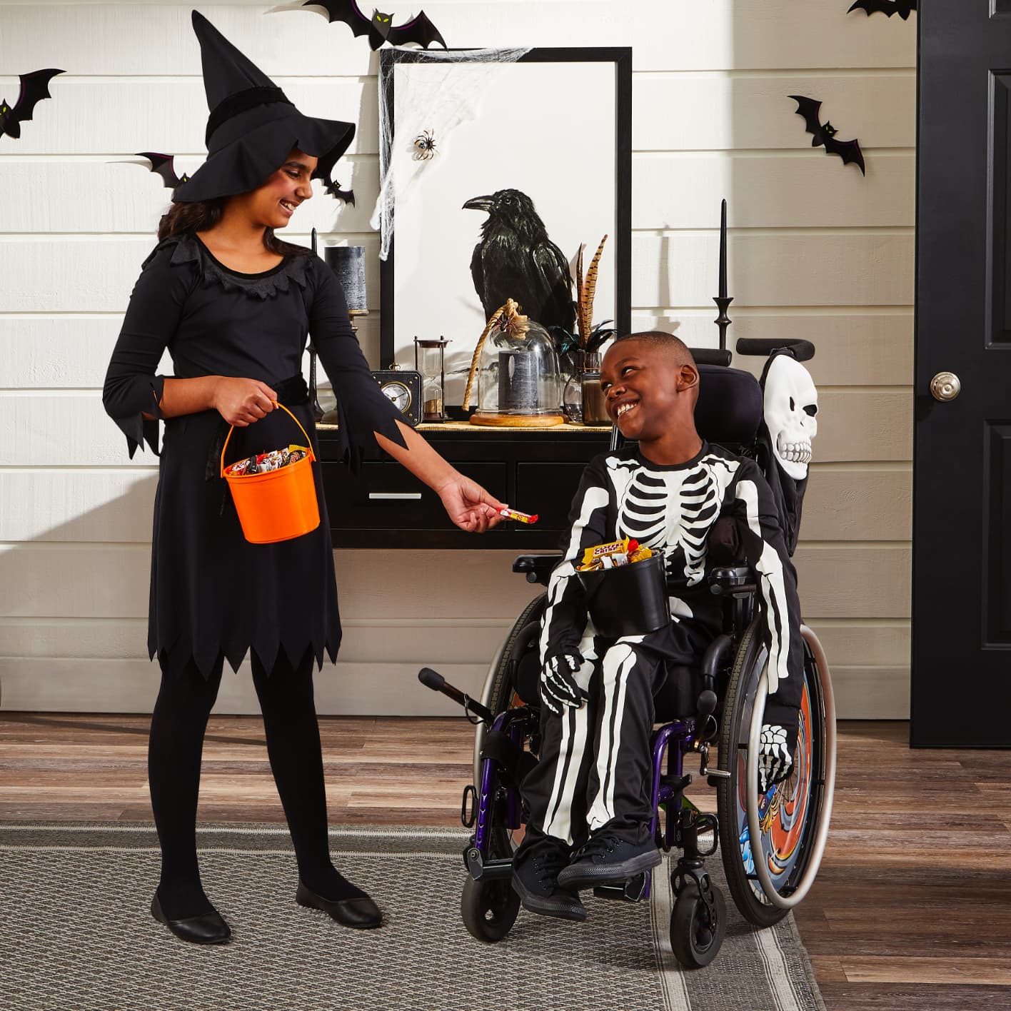 Girl dressed as a witch handing candy to a boy in wheelchair dressed as a skeleton.