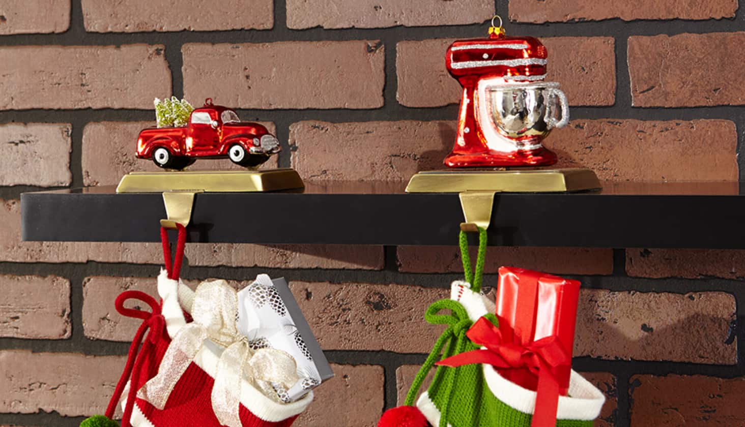 Two stockings hanging on a fireplace with stocking holders decorated with a red truck and stand mixer decorations.