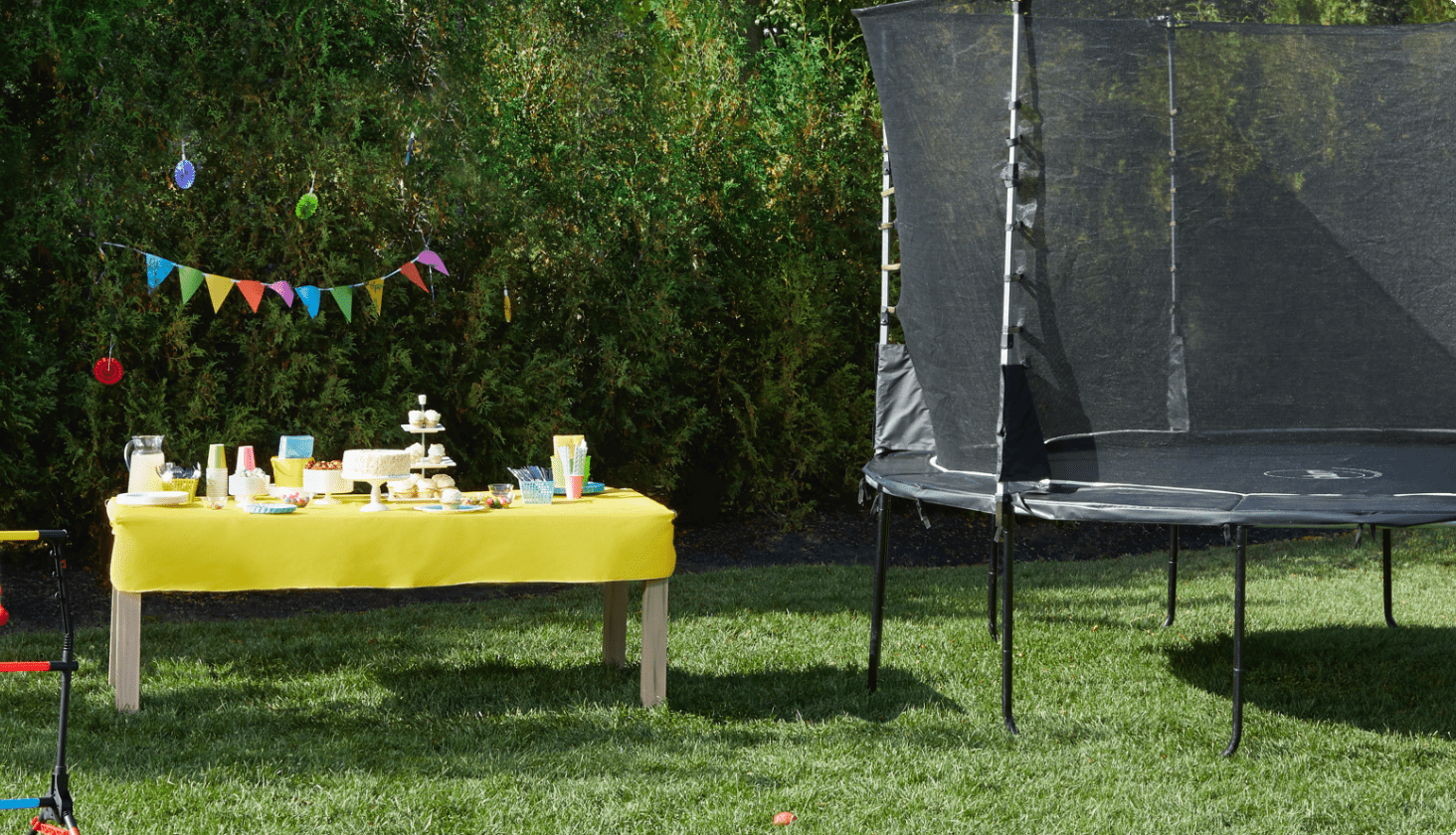 Birthday party table set up with yellow table cloth, birthday cake and party cups beside a trampoline in a backyard.