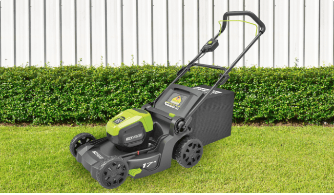 A 17-inch Greenworks 60-volt Cordless Lawn Mower stands on green grass in front of a low hedge and a white fence.