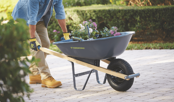A person wearing yellow gardening gloves pushes a Yardworks Steel Wheelbarrow filled with flowers across patio stones in a backyard.