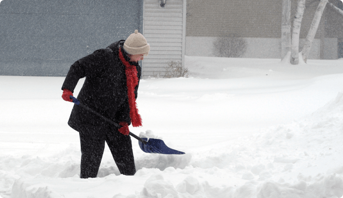 A person wearing a black coat, hat, and gloves uses a shovel to clear snow.