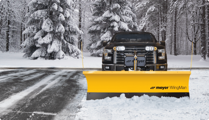 A black pickup truck with a yellow snow plow blade mounted to its front moves snow off a driveway. 