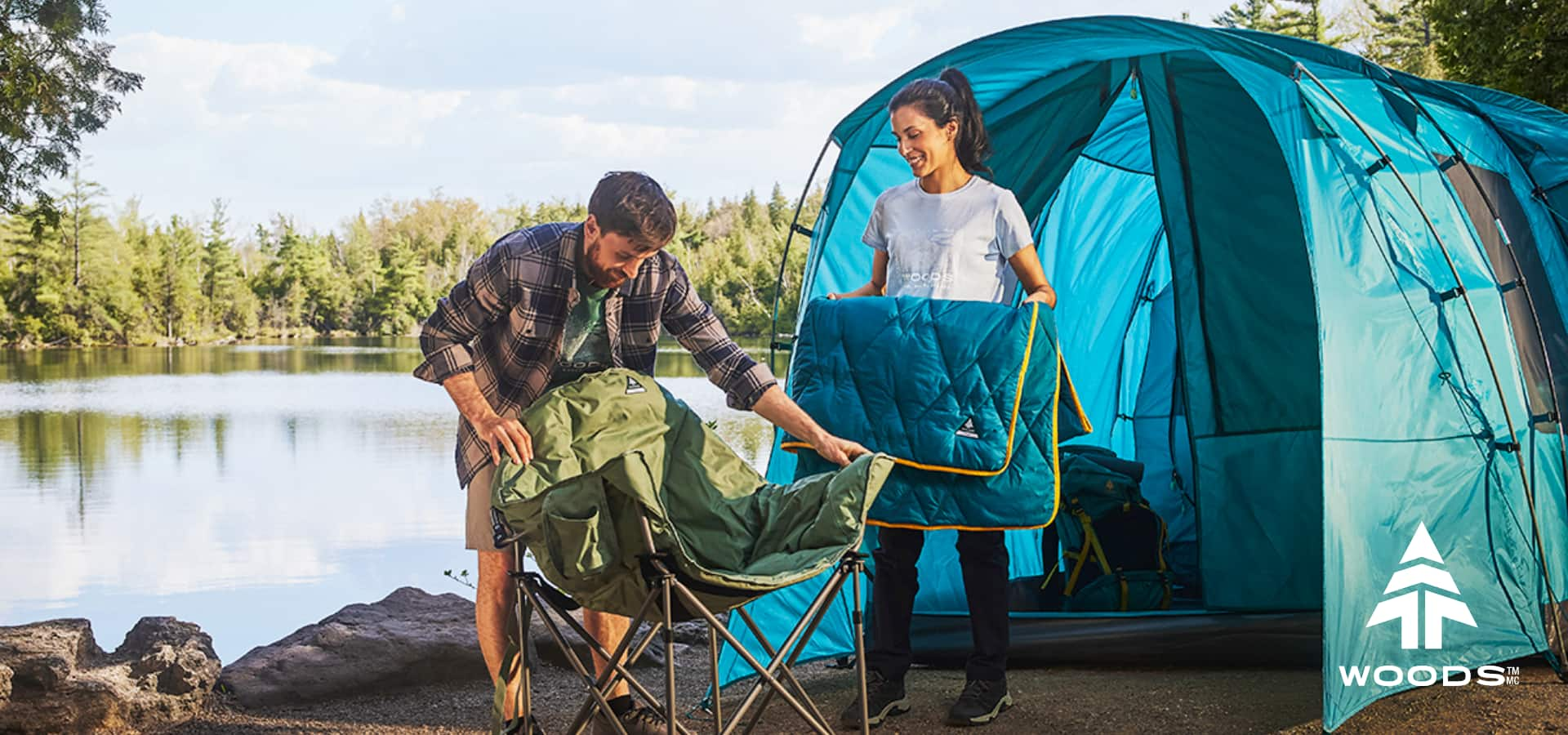 A couple happily setting up camping chair next to their tent, near a lake.